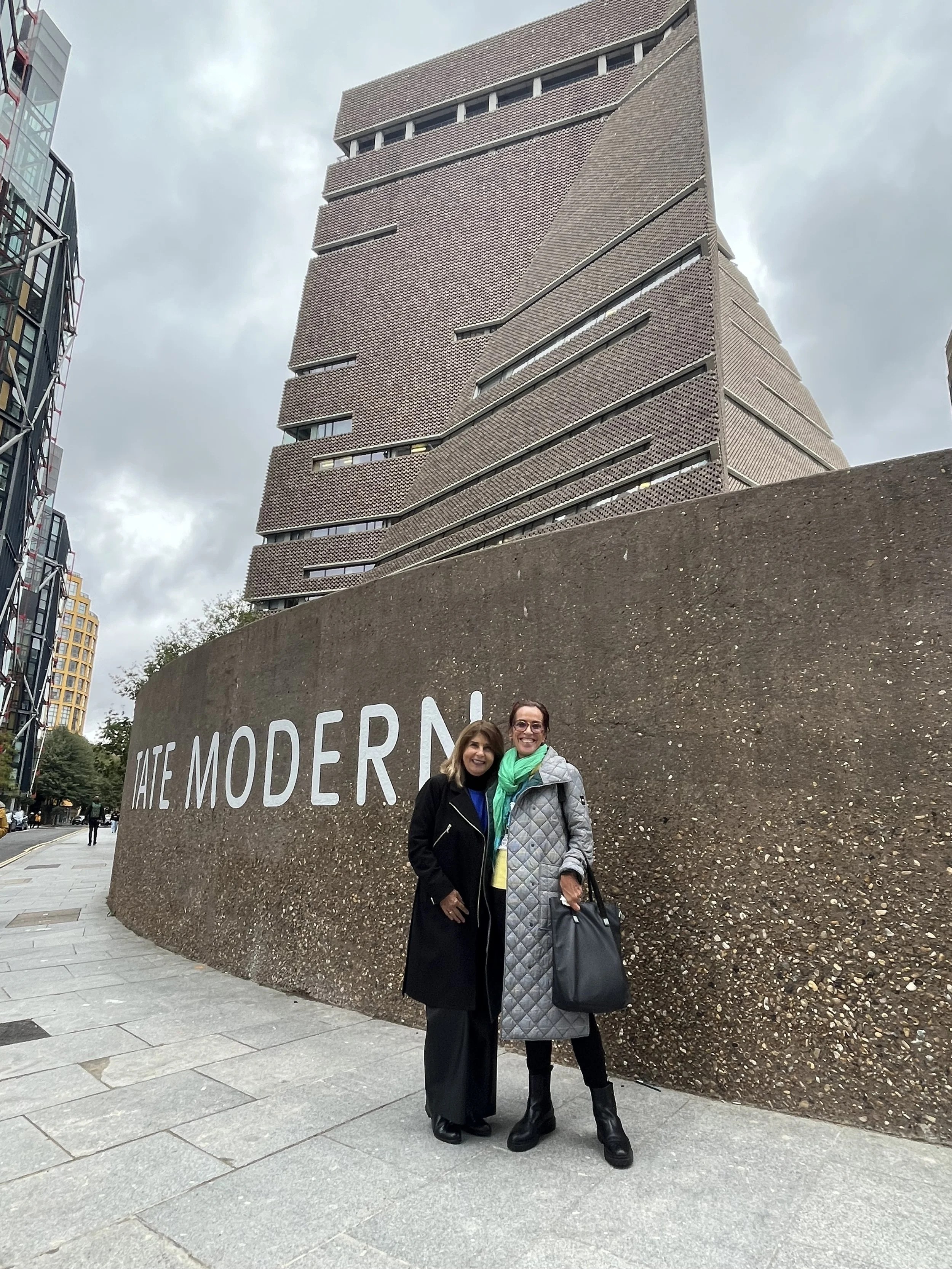 Two women standing in front of a curved wall with the words 'The Modern' written on it, overlooking a modern skyscraper with an unusual architectural design, with other tall buildings in the background on a cloudy day.