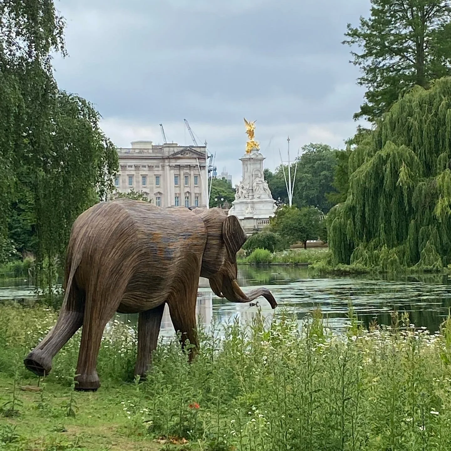 Wooden elephant sculpture near a river with greenery, a tall white monument with a golden statue on top, and buildings in the background.