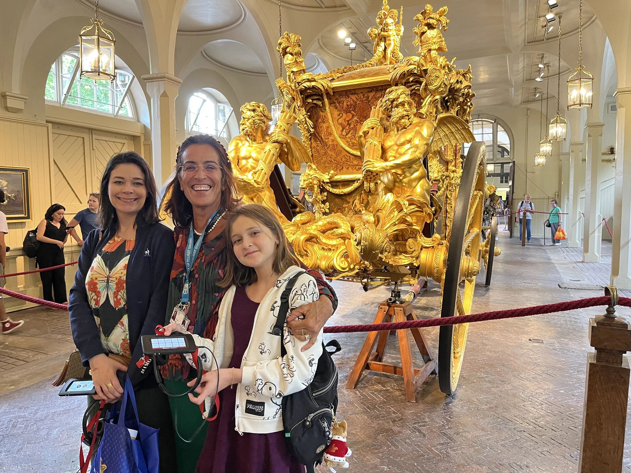 Three women and a girl smiling at a museum, with a large ornate golden carriage in the background. The carriage has intricate details and is displayed behind a red rope barrier.