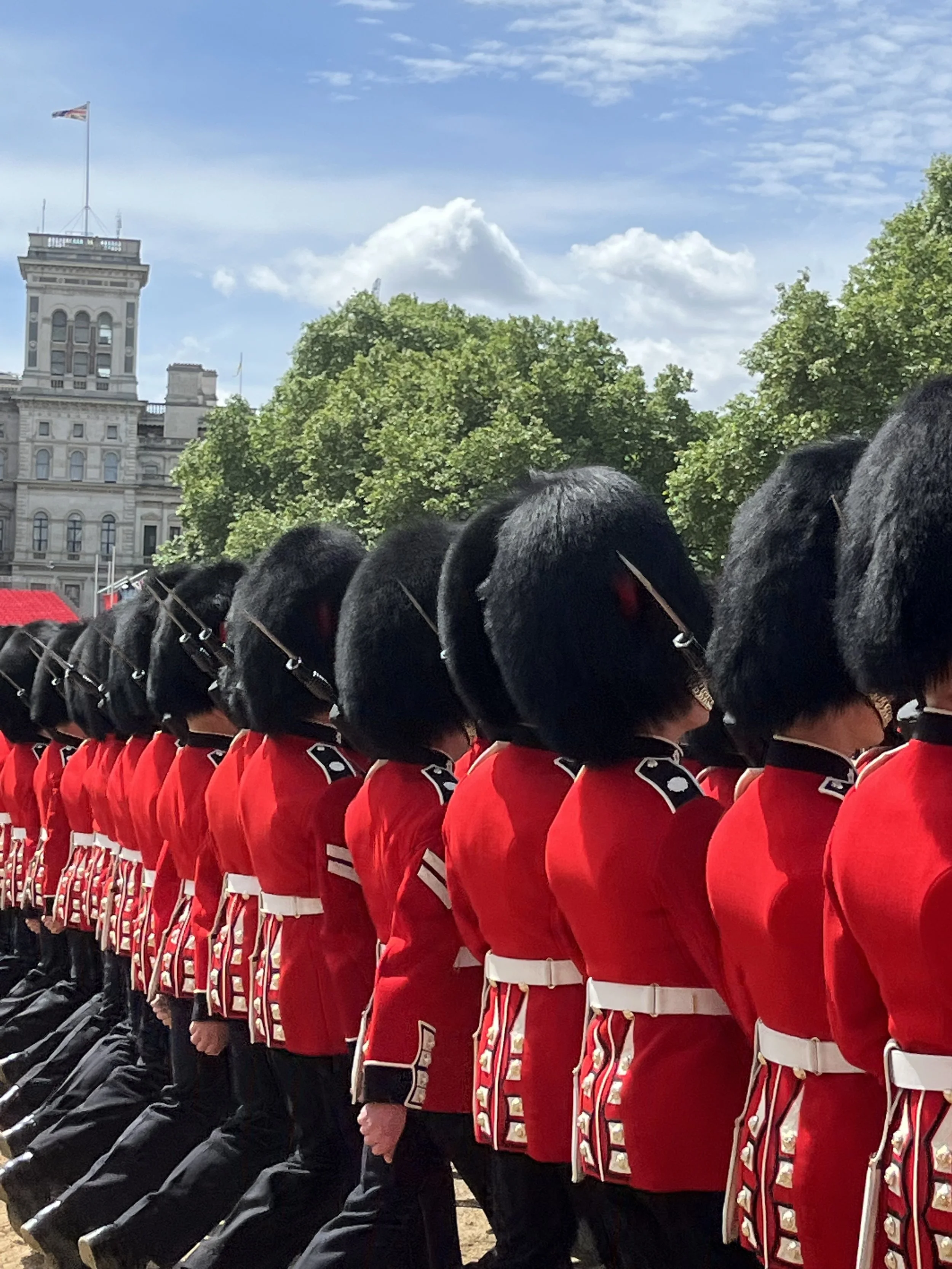 Line of soldiers in red uniforms and black bearskin hats during a parade in an outdoor urban area with historic buildings and green trees, under a partly cloudy sky.