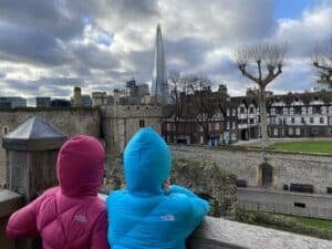 Two children in pink and blue jackets sitting on a wooden fence, looking at the London skyline including The Shard, on a cloudy day.