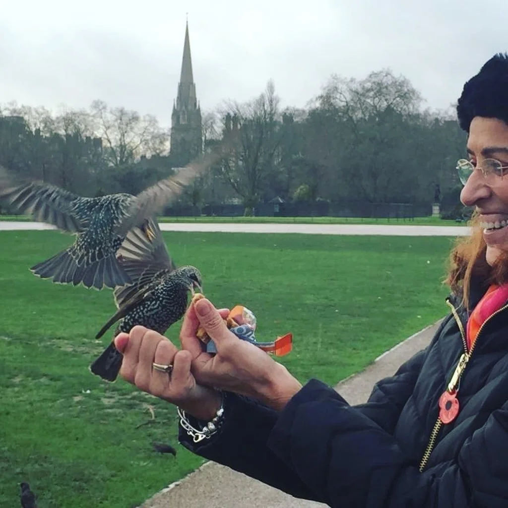 A woman holding a small bird on her hand on a park path with trees and a church spire in the background, during overcast weather.