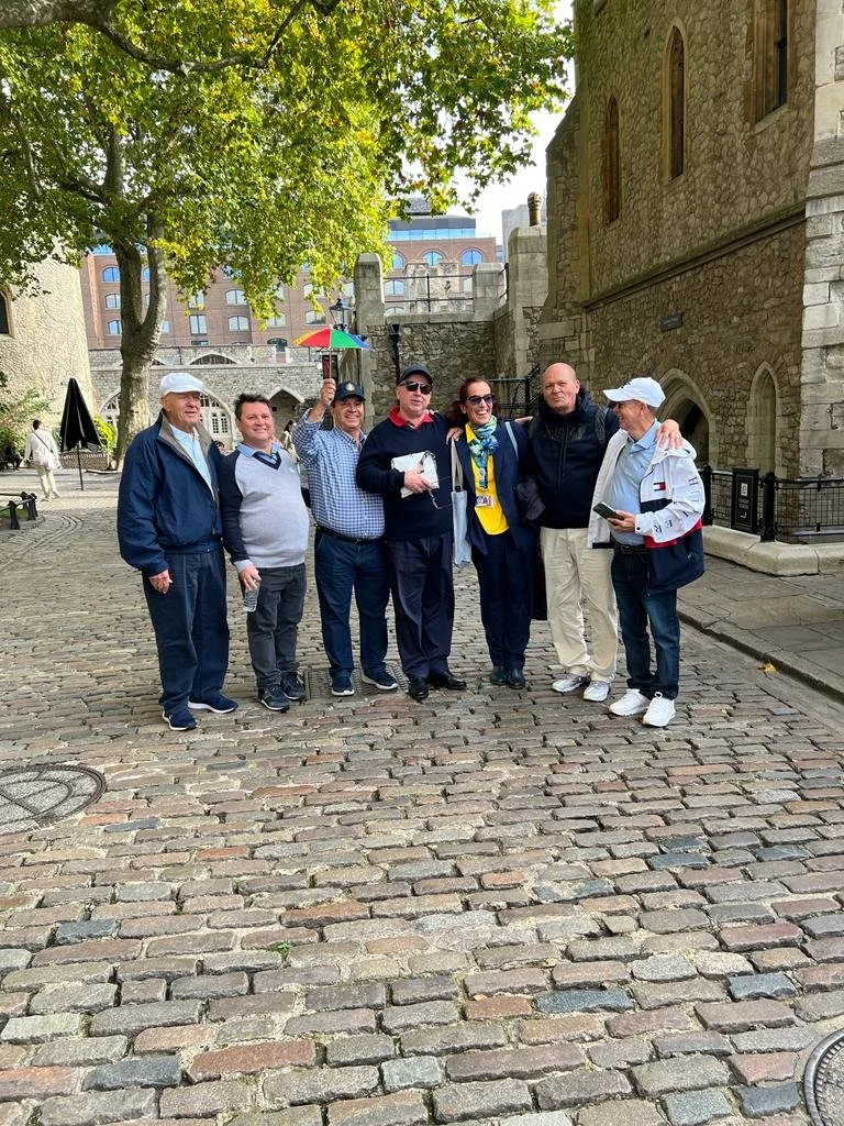 A group of seven people standing on a cobblestone street in front of a historic stone building with a crenellated wall, surrounded by trees, with some modern buildings in the background, during the daytime.