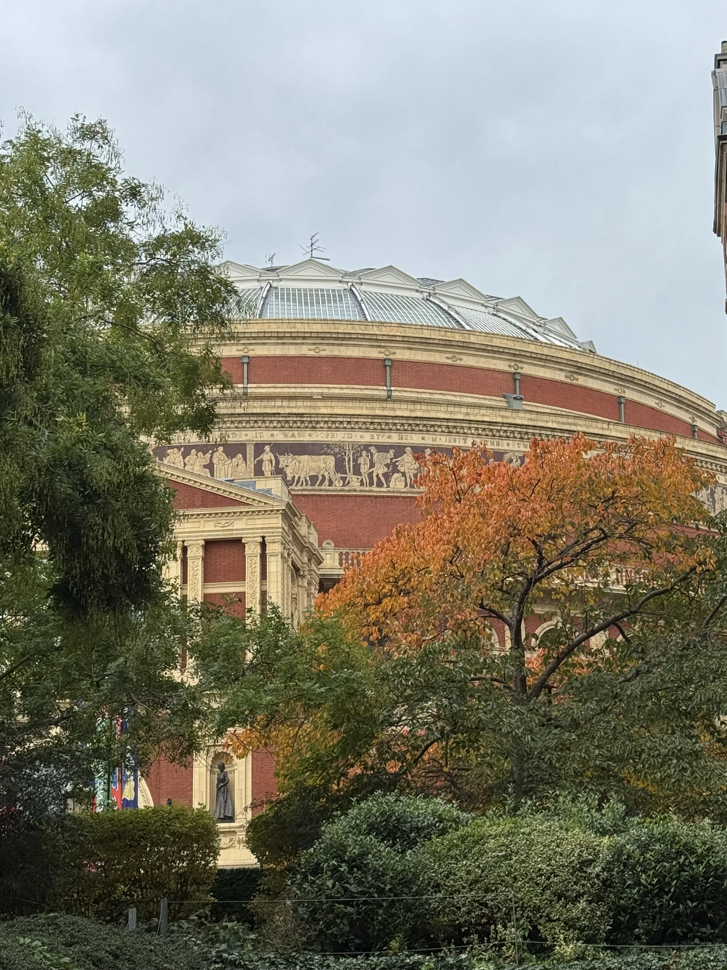 Historic building with detailed architecture and a frieze, partly obscured by trees with green and orange leaves, under an overcast sky.
