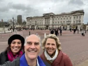 Three people taking a selfie in front of a large historic building on a cloudy day.