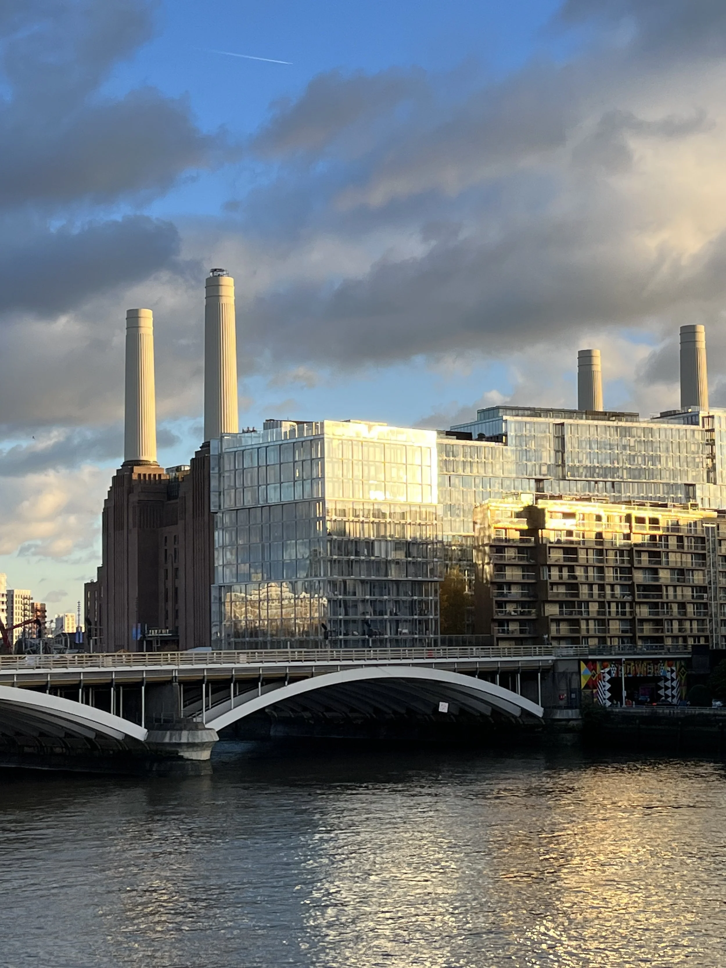 A city skyline with modern glass buildings reflecting sunlight, a bridge over a river, and four tall smokestacks against a partly cloudy sky at sunset.