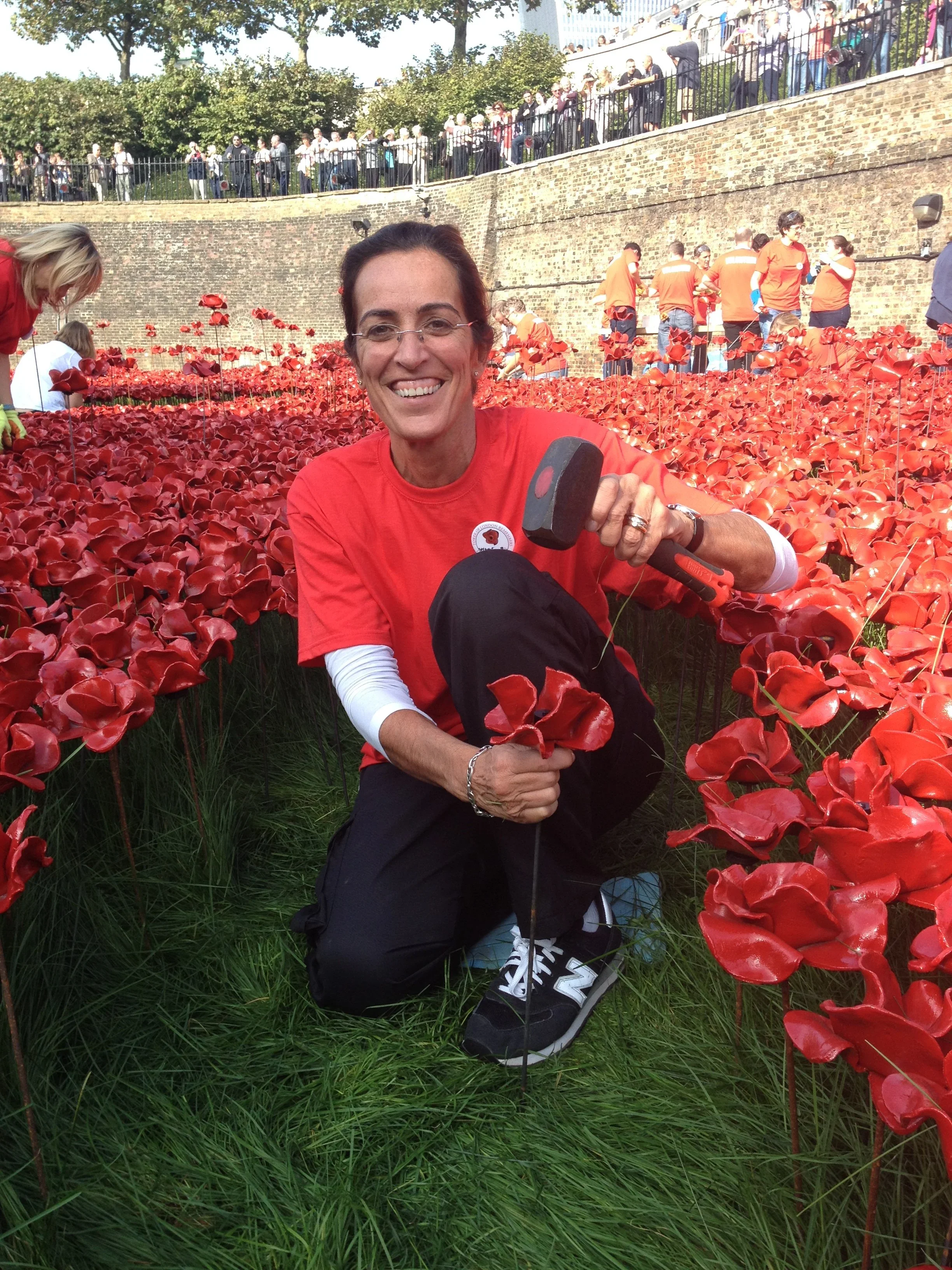 A smiling woman kneeling among red artificial flowers in a garden, holding a small hammer and a flower, with a crowd and a brick wall in the background.