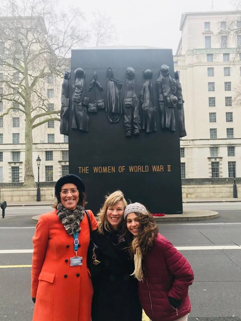 Three women standing in front of the Women of World War II memorial in Washington D.C., smiling. The memorial features a black background with metallic sculptures of women and the inscription 'The Women of World War II'.