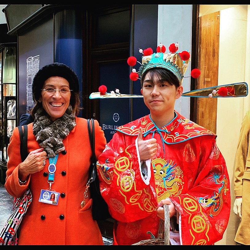 A woman wearing a dark hat, glasses, orange coat, and a furry scarf, smiling next to a young man in traditional Chinese attire, including a bright red robe with intricate embroidery and a decorative headpiece with red pompoms, giving a thumbs up.
