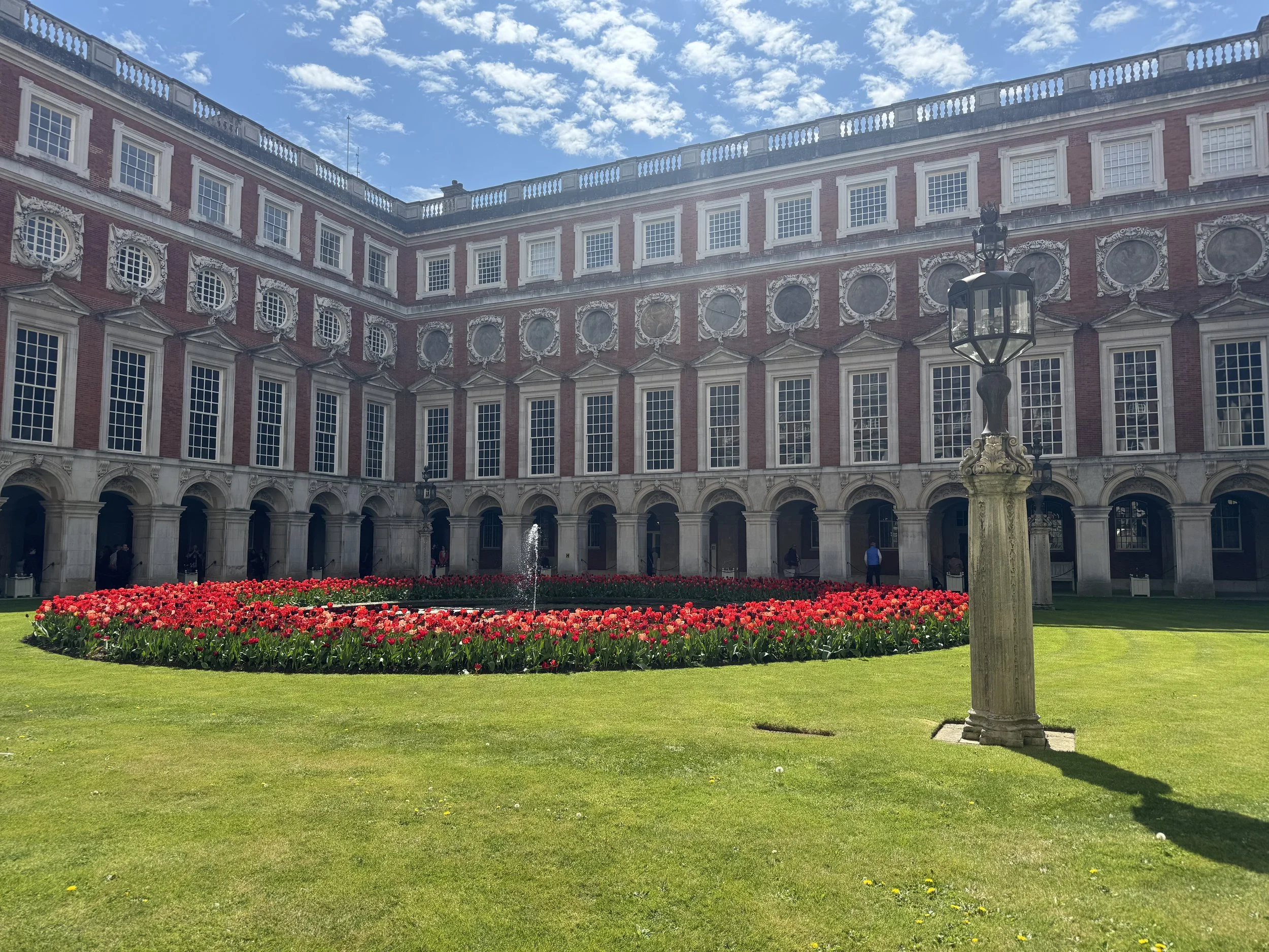 A courtyard within Hampton Court a well-maintained green lawn, a circular flower bed with red tulips, a fountain in the center, and a tall vintage lamp post. In the background, there is a large historic building with multiple windows and an ornate ex
