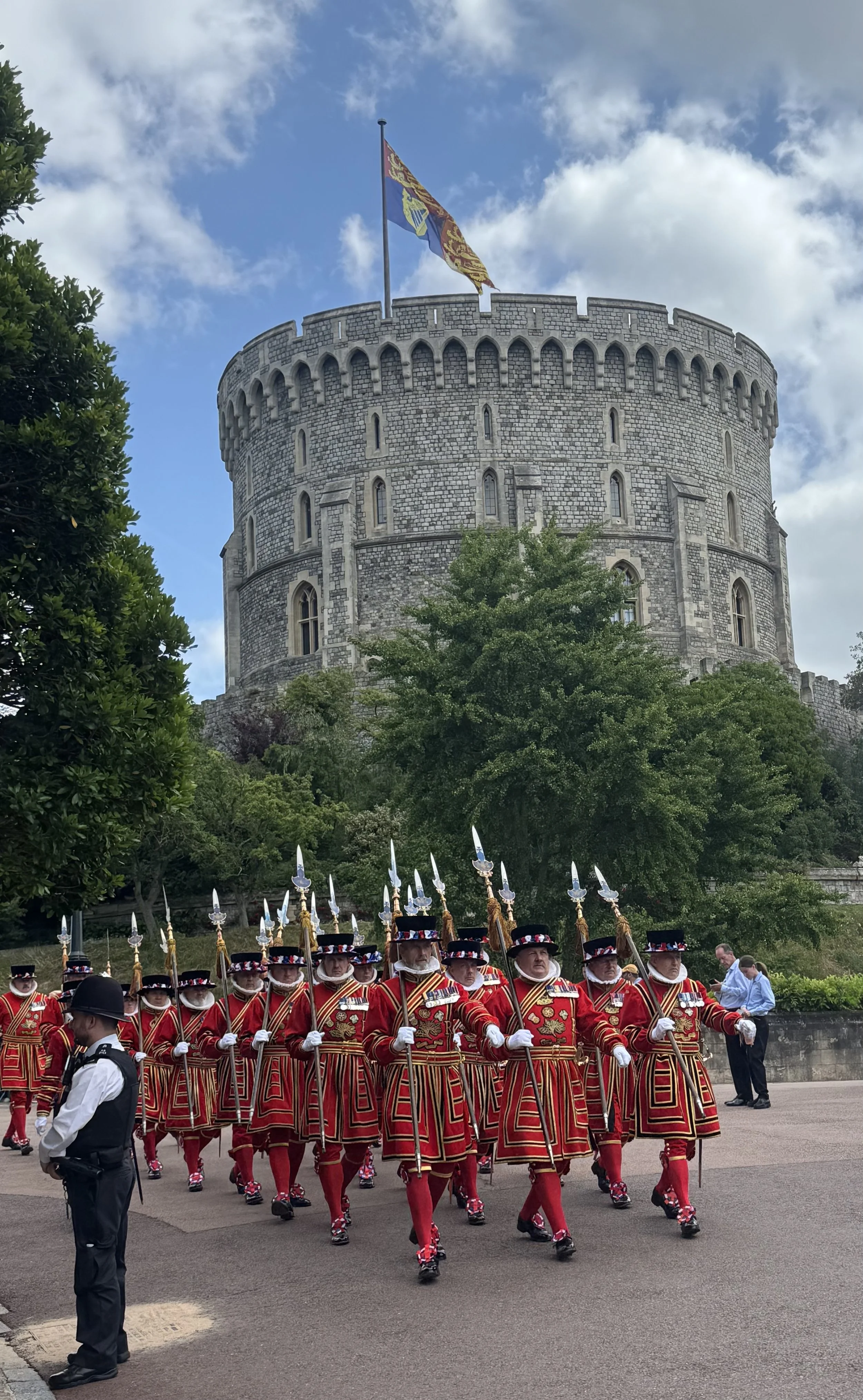A group of the Queen's Guard soldiers in red uniforms and bearskin hats marching in formation near Edinburgh Castle. The castle is a large stone fortress with a flag flying at the top. The sky is partly cloudy.