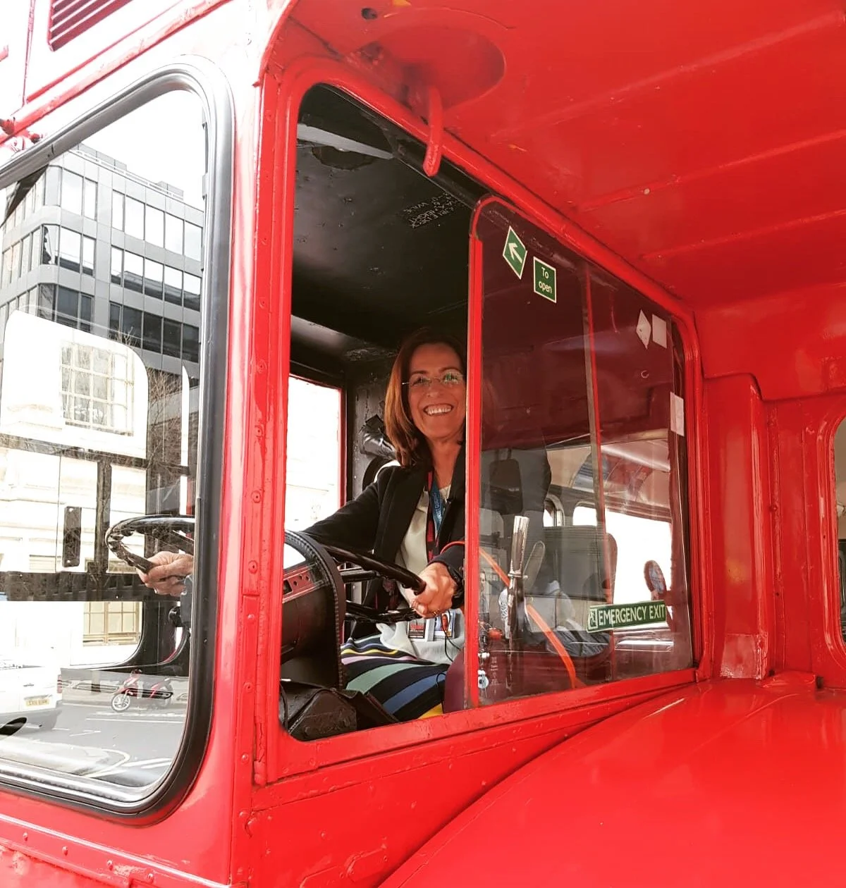 A woman smiling while sitting in the driver's seat of a red vintage vehicle, holding the steering wheel, with a city building visible outside the window.