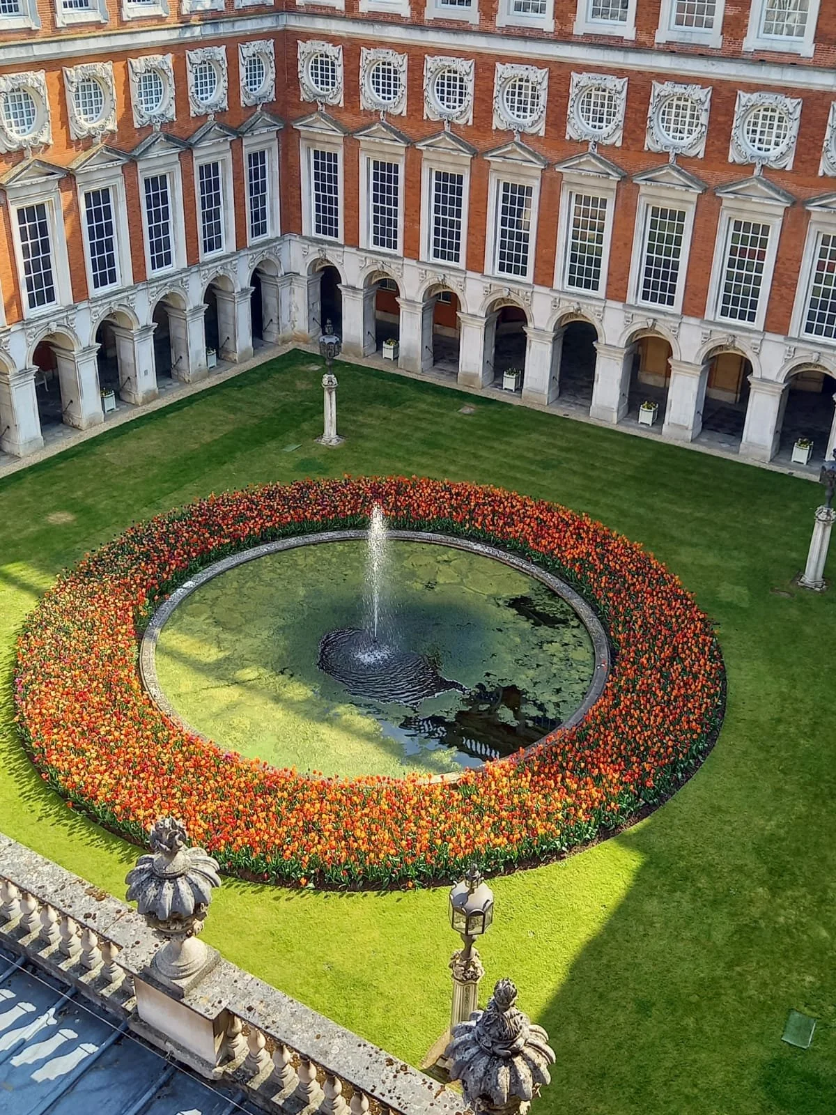 A courtyard with a circular pond surrounded by a vibrant orange flower bed, a fountain in the center, and a historical building with arched windows and decorative stonework.
