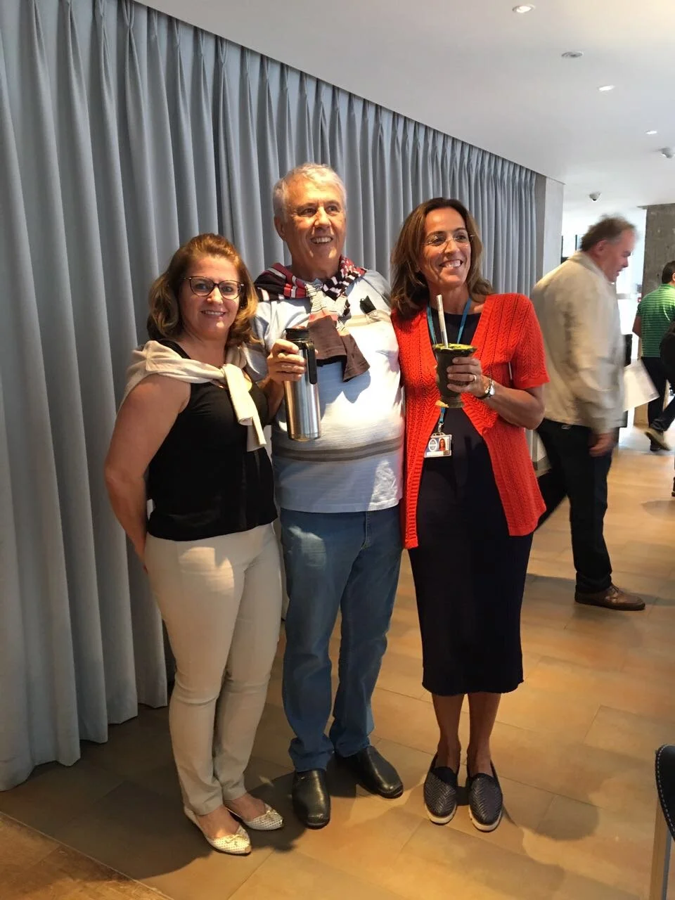 Three people standing together at an indoor social event, smiling, with two women and one man. The woman on the left has shoulder-length hair, glasses, a black top, and beige pants. The man in the middle has short gray hair, a striped shirt, and is h