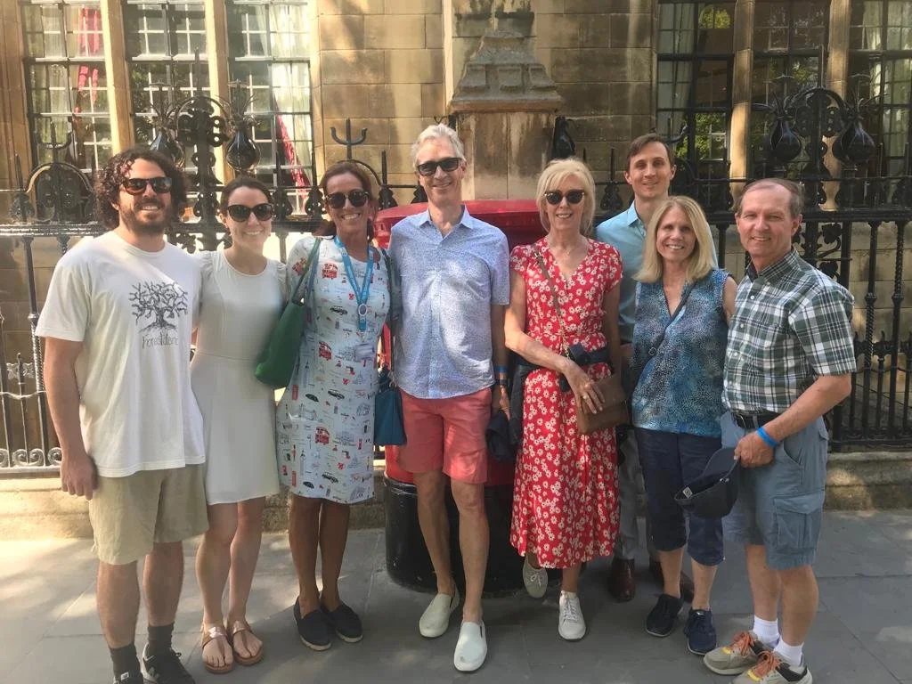 Group of nine people standing outdoors in front of a historic stone building with iron fence, mostly smiling.