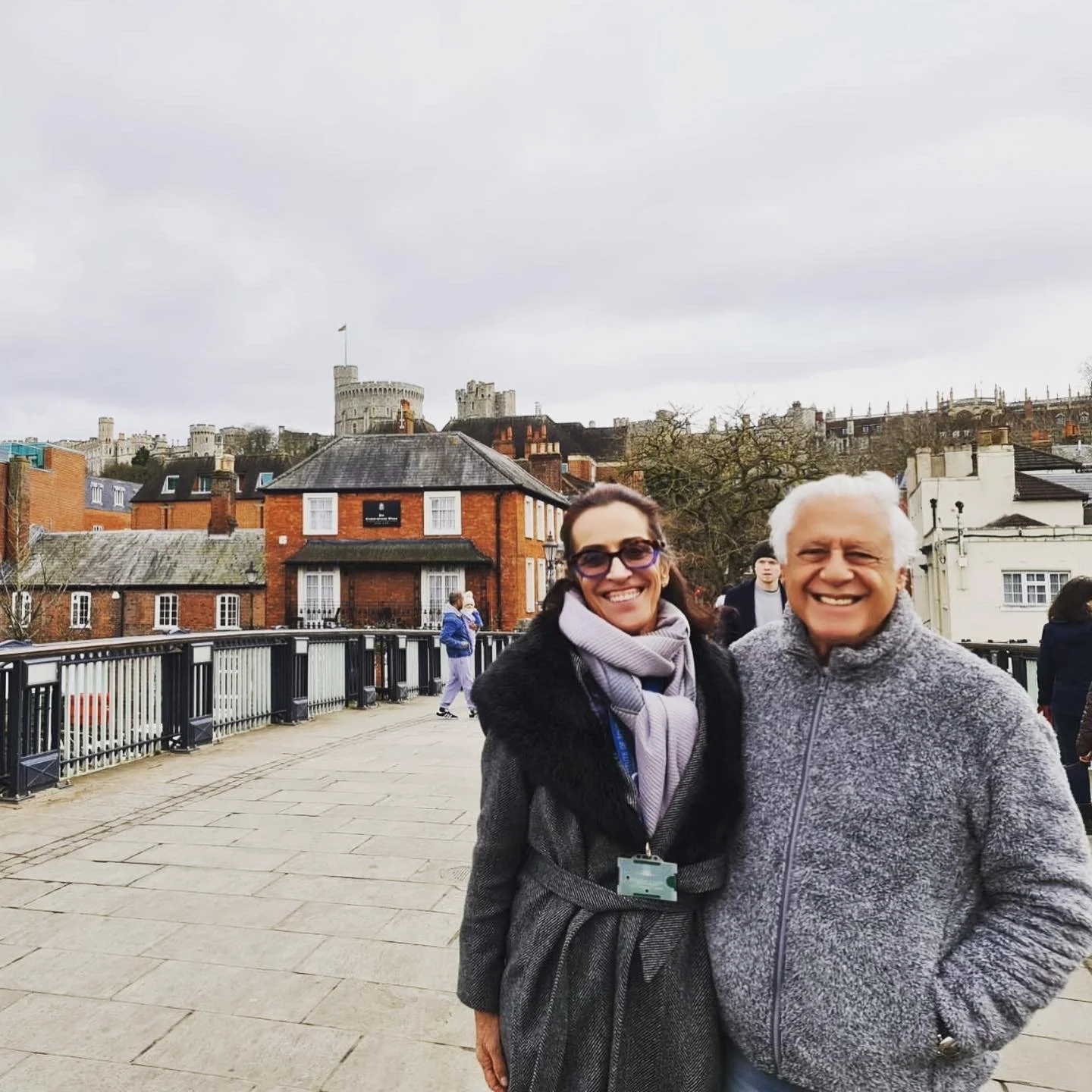 Smiling woman and older man posing on a bridge with a view of a city and a castle in the background.