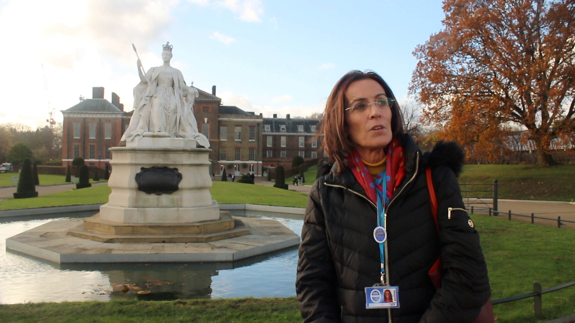 A woman with glasses and a black jacket standing in front of a fountain with a statue of a queen holding a scepter, with historic buildings and autumn trees in the background.