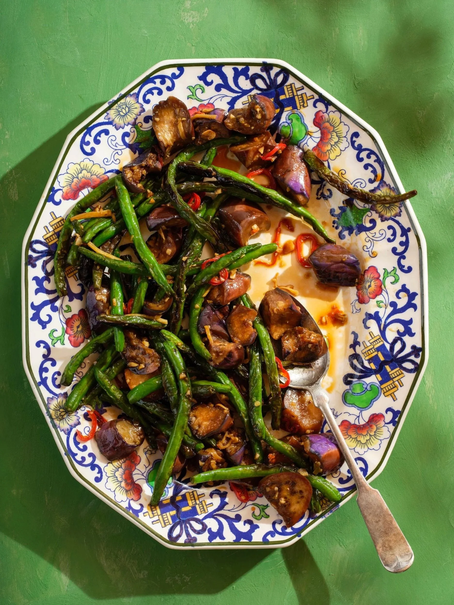 A plate of sautéed green beans on a decorative platter with a spoon resting on the side of the plate. Mixed with garlic, hot chiles and mushrooms.