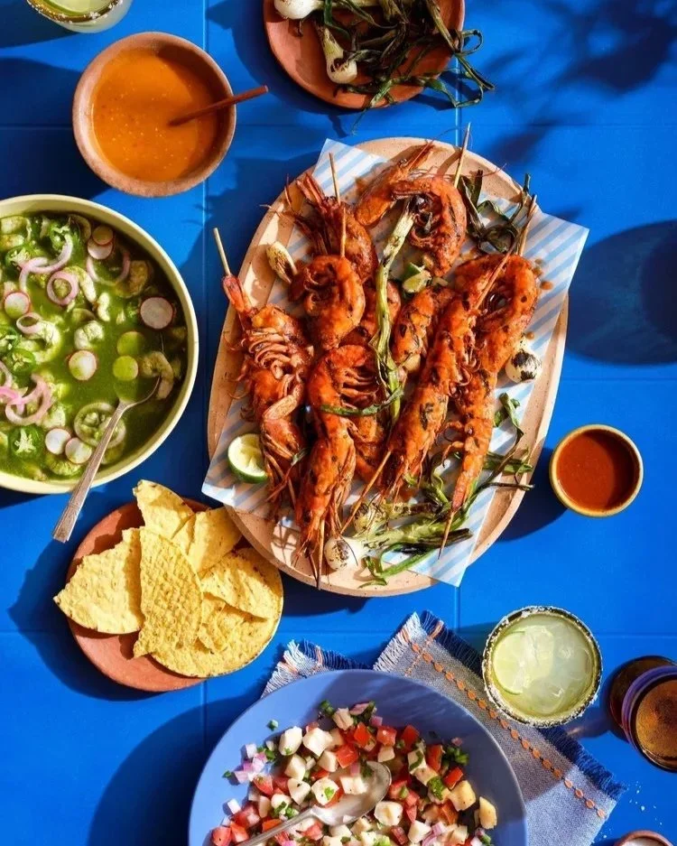 a dish of grilled shrimp sit on a terra cotta platter surround by chips, salsa, pico de gallo on a blue tabletop