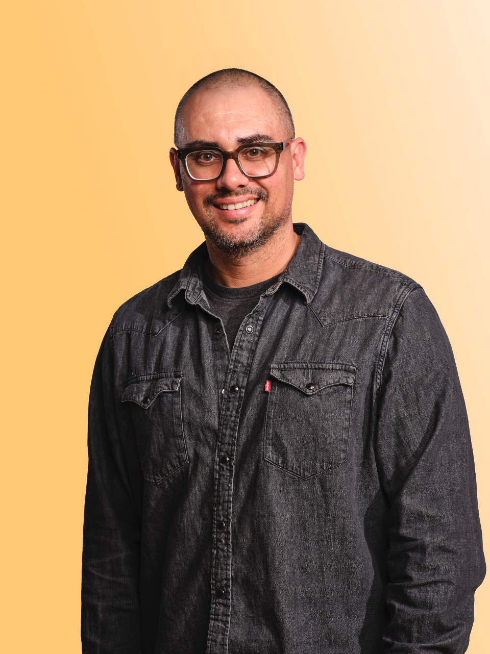 Male of asian or hispanic descent, wearing a black levis denim shirt and brown glasses on a yellow background.