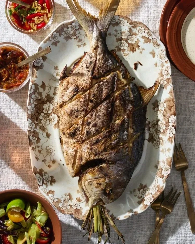 A whole fried fish on a white decorative platter. Shot overhead with sides of hot sauce, limes and warm summer light.