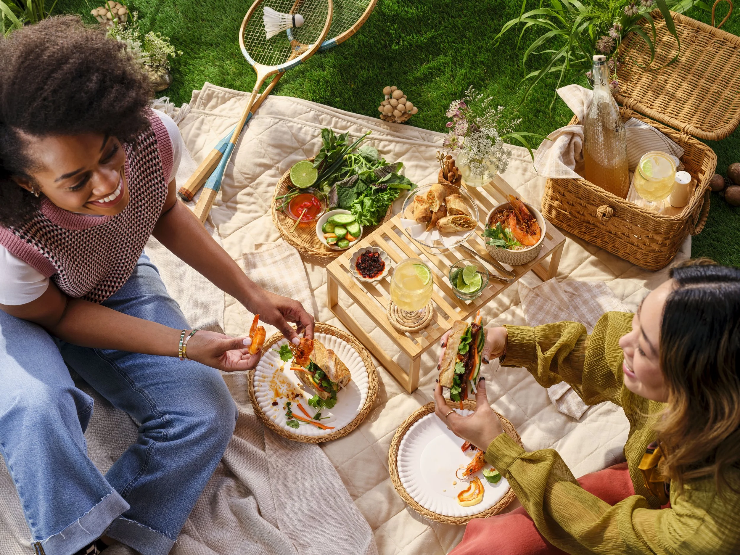 Two women sitting on a blanket having a picnic outside in the summer.  They are eating bahn mi, chilled shrimp, cold drinks.  The blanket they are sitting on is in the grass with tennis rackets and a picnic basket.