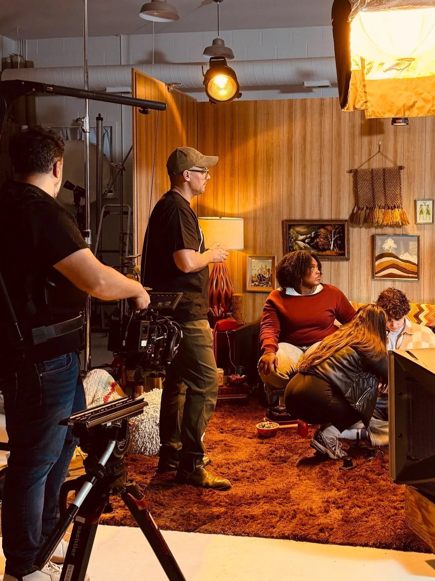 Behind the scenes of a video set. The director, dp and talent are on set waiting for direction. The scene is a midwest 70s style living room with shag carpet and wood paneling on the wall.