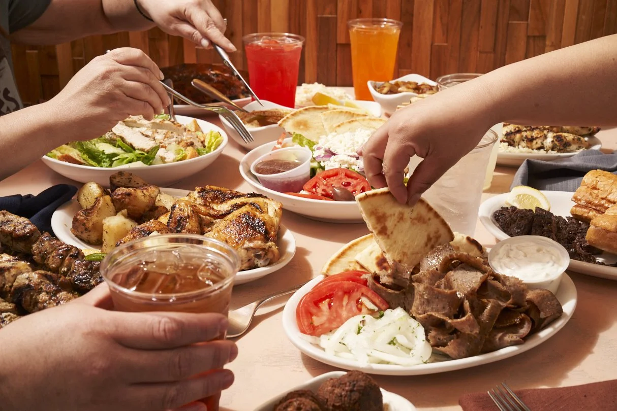 A dinner table full of mediterranean food. Lemon chicken, gyros, pizza, cold sodas and ice tea. Hands are reaching into the frame to grab food and drinks.