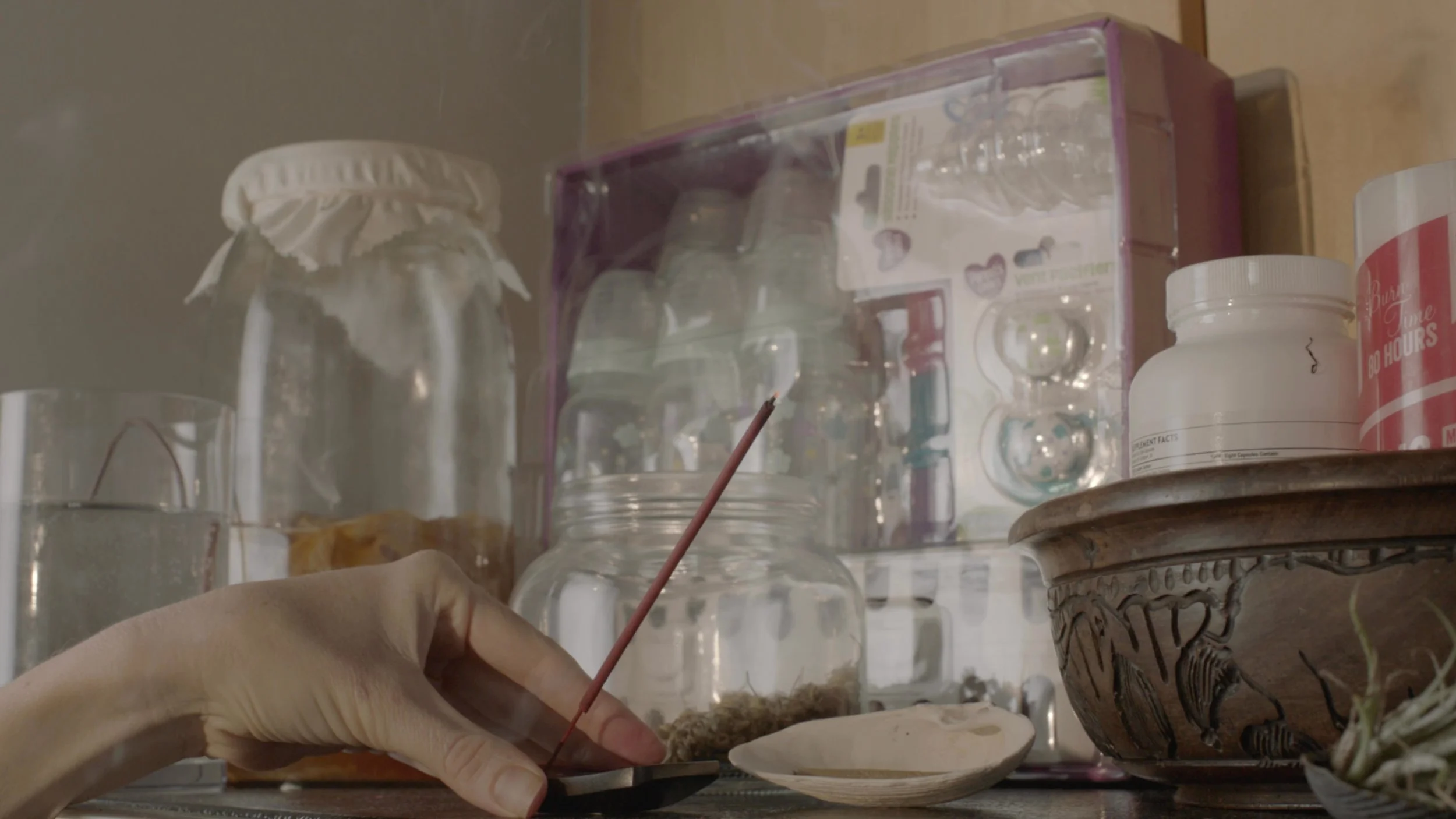 A close-up view of a person’s hand holding a paintbrush over a ceramic bowl, with glass jars, a plastic container, and other miscellaneous objects on a shelf in the background.