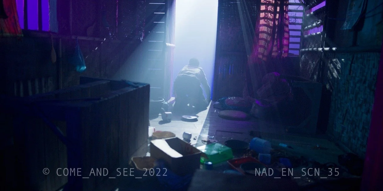 A person kneeling on the floor inside a dimly lit, cluttered room with colorful fabric hanging on the walls, illuminated by bright light coming through a doorway.