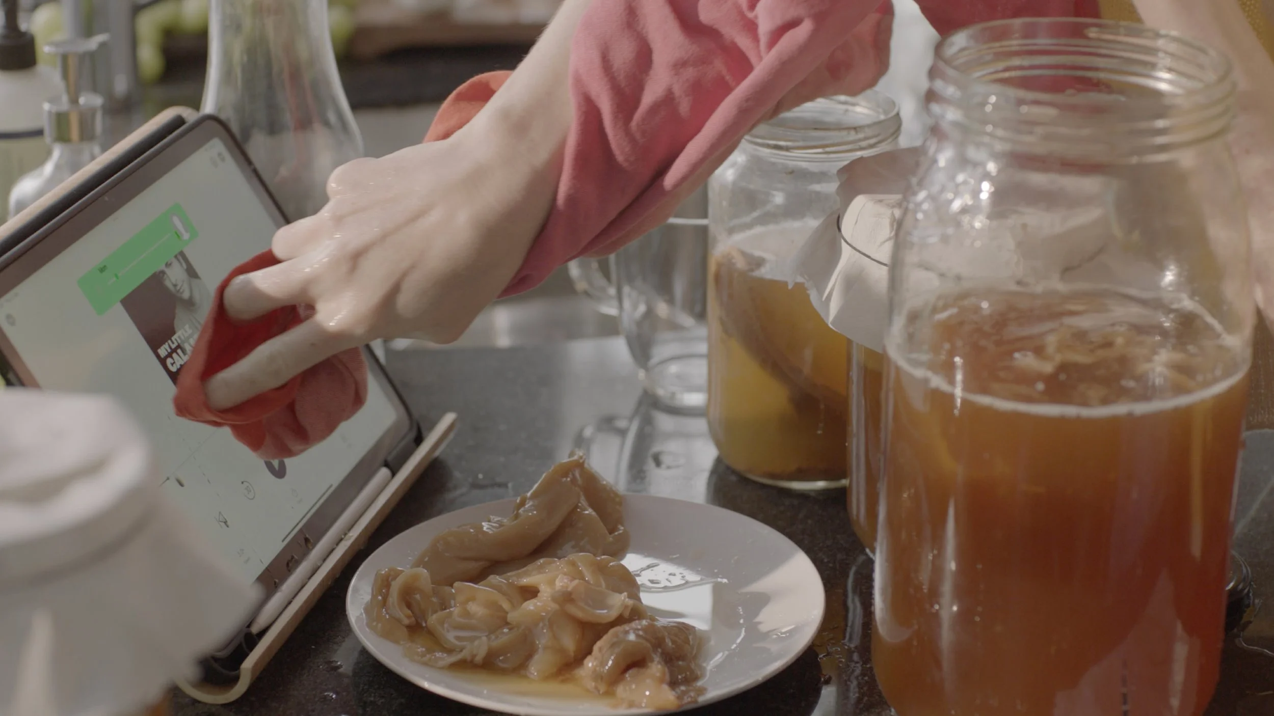 Person cleaning a glass jar with a pink cloth, surrounded by jars, a plate of raw chicken, and drinks on a kitchen counter.
