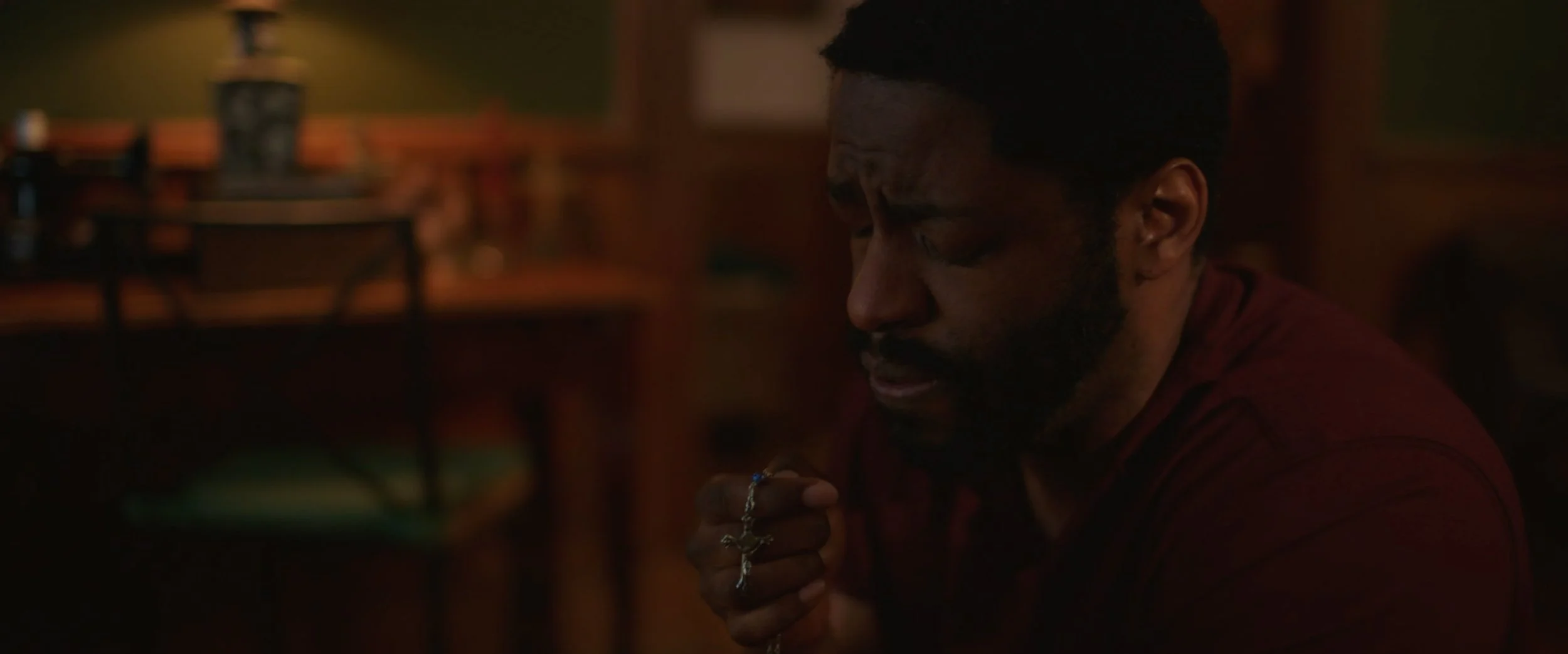 A man with a beard and dark hair, wearing a red shirt, holding a rosary with a cross, eyes closed, appears to be praying in a dimly lit room.