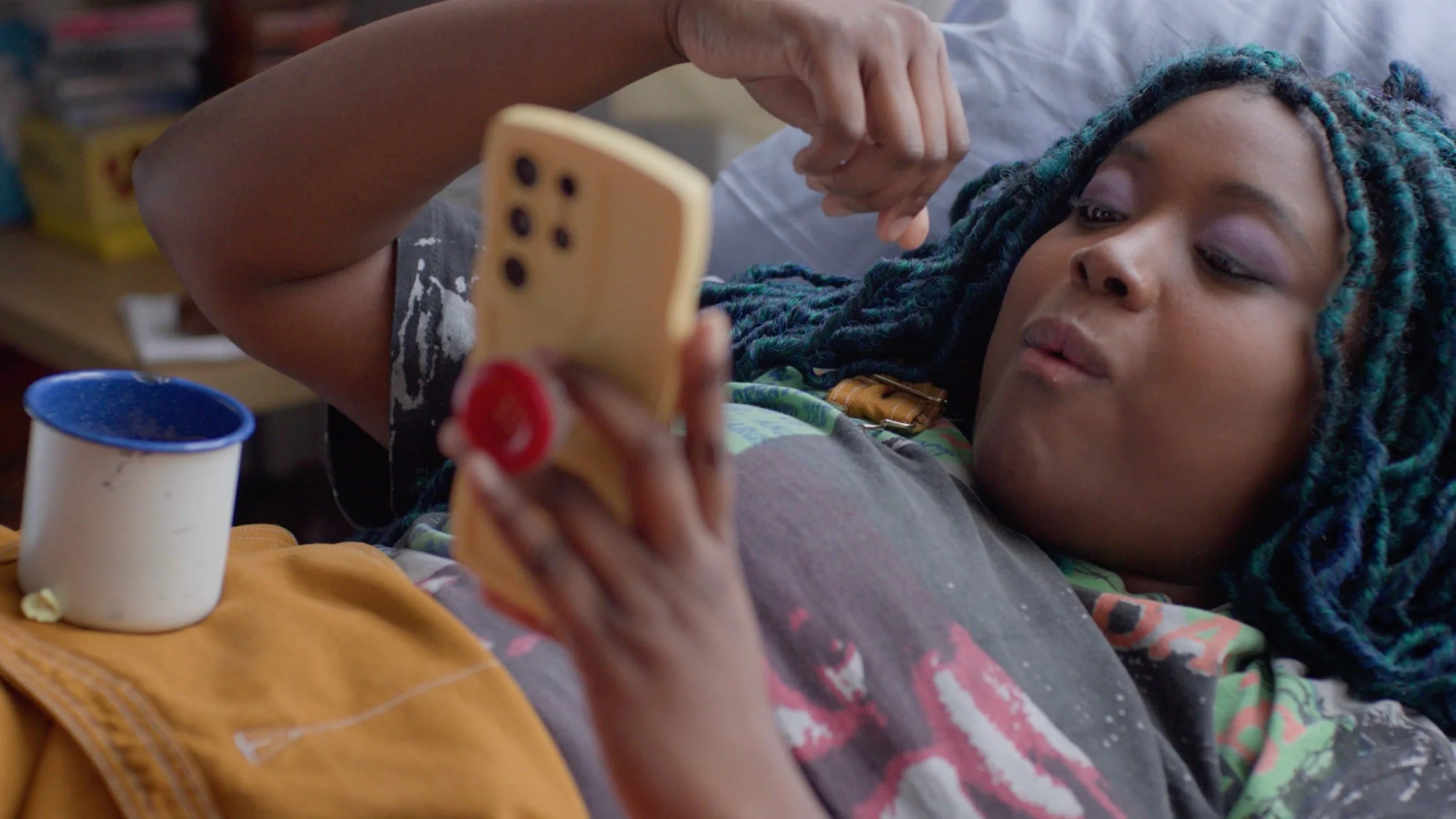 A person with blue and black dreadlocks lying on a bed, looking at a phone in their hand. There is a mug on a yellow blanket in front of them.
