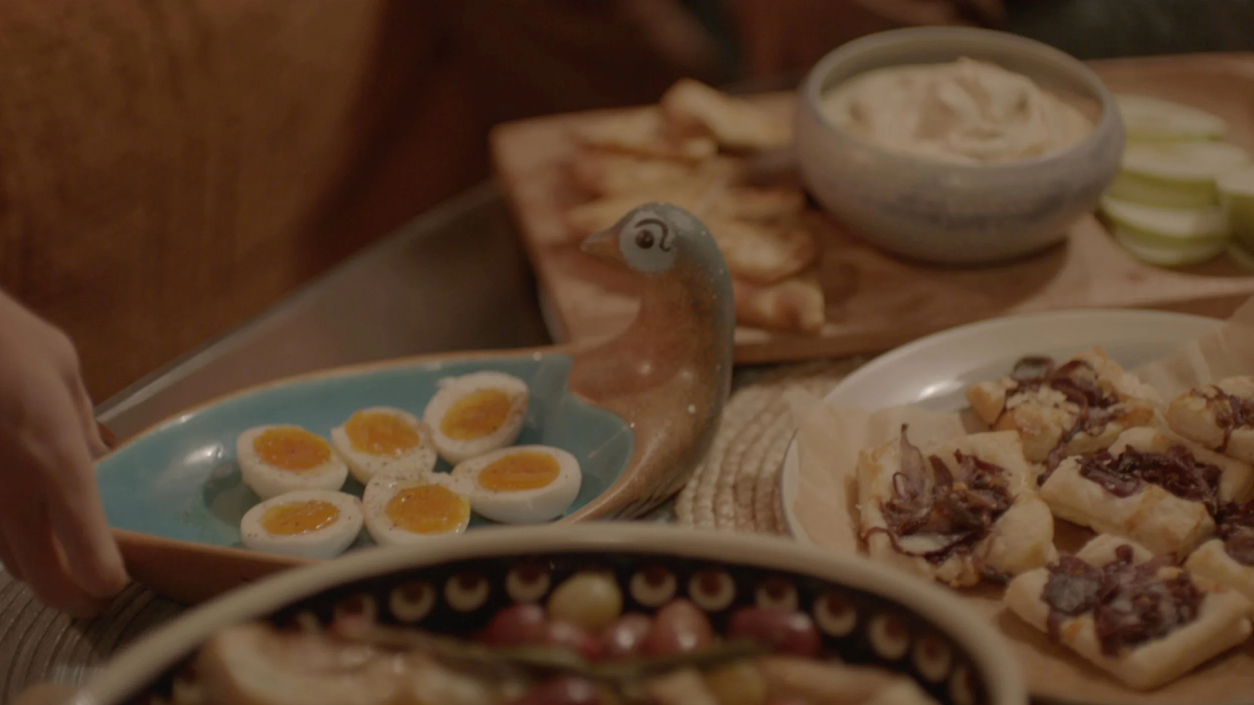 A hand holding a plate of sliced soft-boiled eggs with a yellow yolk, surrounded by other plates of assorted baked and sliced foods on a wooden table.