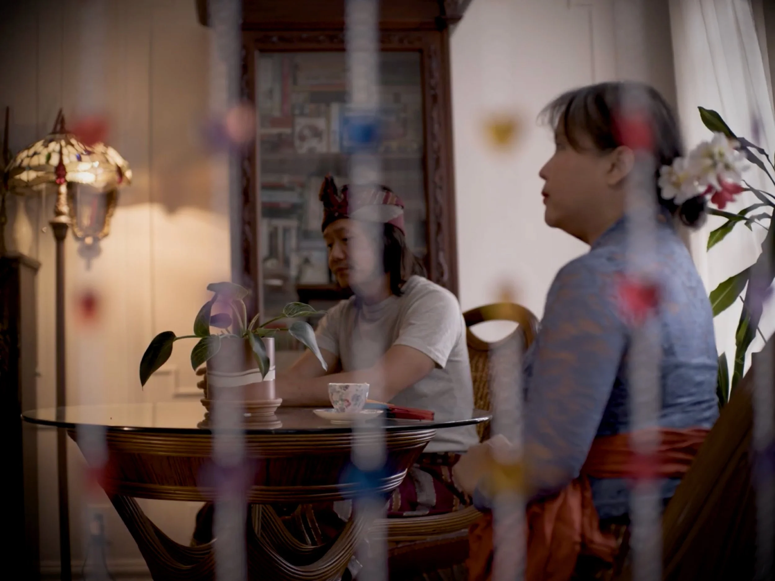 A person with a bun hairstyle and floral hair accessory sits at a dining table with another person with long hair wearing a patterned shirt. The scene is viewed behind a decorative glass table with floral arrangements, a lamp, and a background of a b