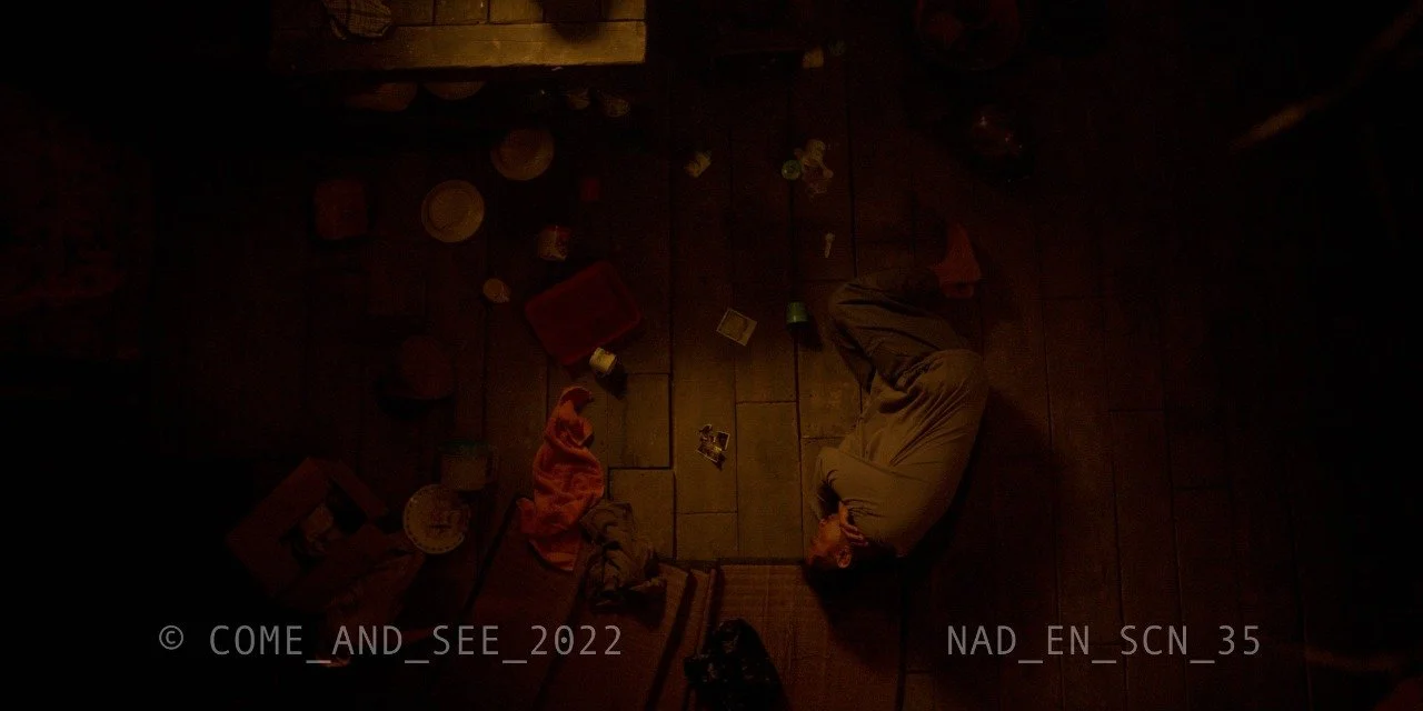 A person sleeping curled up on a wooden floor surrounded by scattered toys and household items in dim lighting.