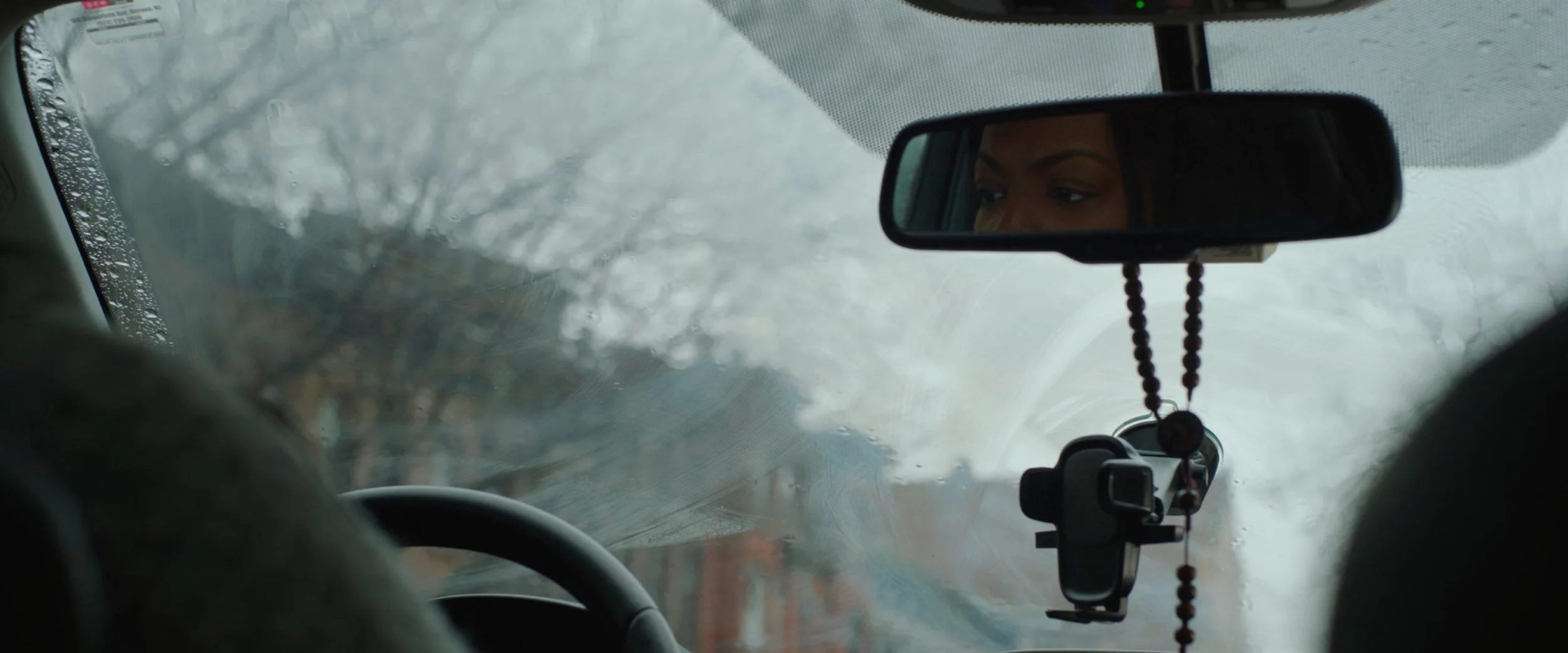 A person driving a car in rainy weather, seen through the windshield with water droplets, and looking into the rearview mirror where their face is partially visible.