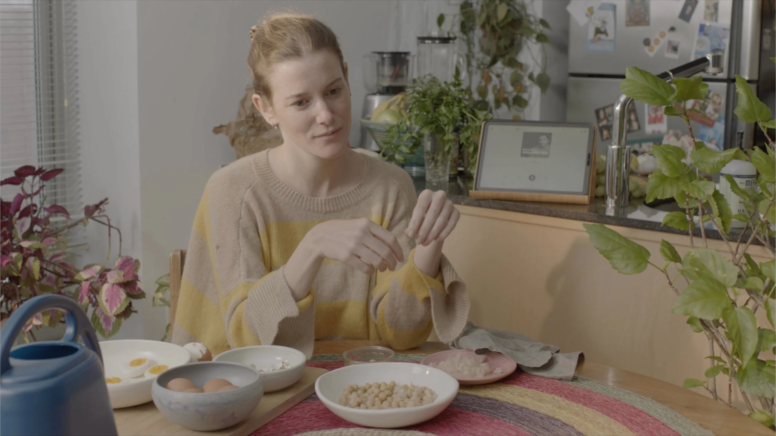 A woman with red hair and a beige and yellow sweater sitting at a kitchen table with bowls of food and ingredients, surrounded by plants and kitchen appliances.