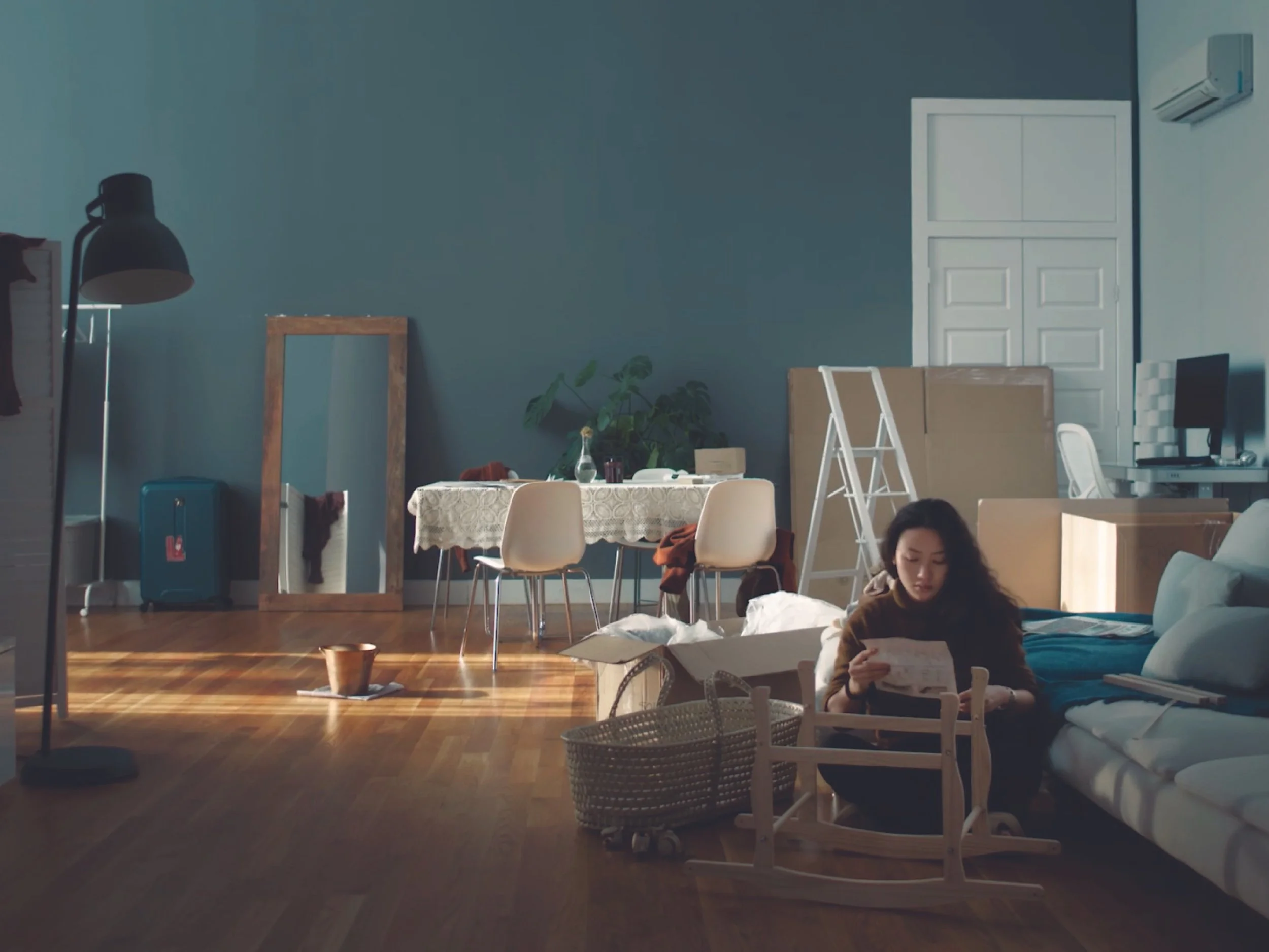 A cozy indoor workspace with a dark blue wall, a white air conditioning unit, a white door, a table with a lace tablecloth, various chairs, plants, and a person sitting on the floor reading a book.