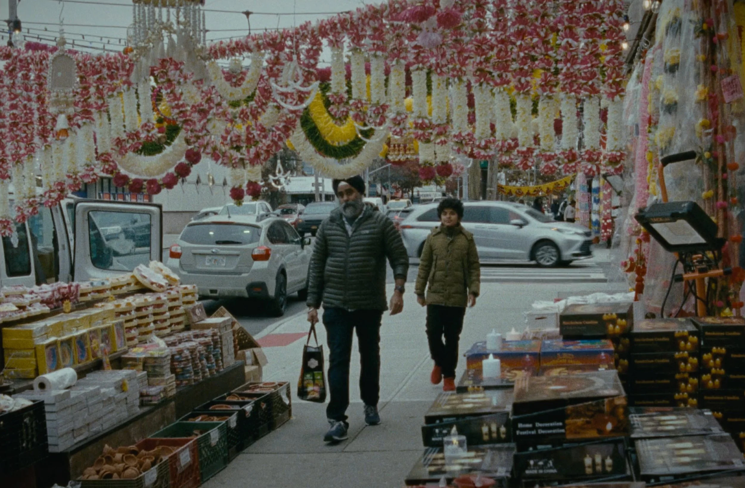 Two men walk through a street market decorated with pink and white floral garlands and ornaments, with cars parked and people in the background.