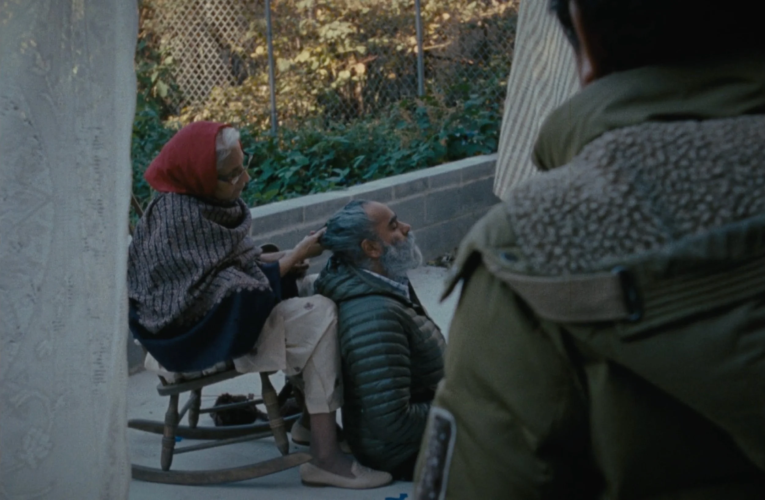 Three homeless individuals sitting on a bench outdoors, with one woman wearing a red hoodie and glasses, a man with a beard wearing a striped puffer jacket, and another woman with long dark hair, in front of a chain-link fence and foliage.