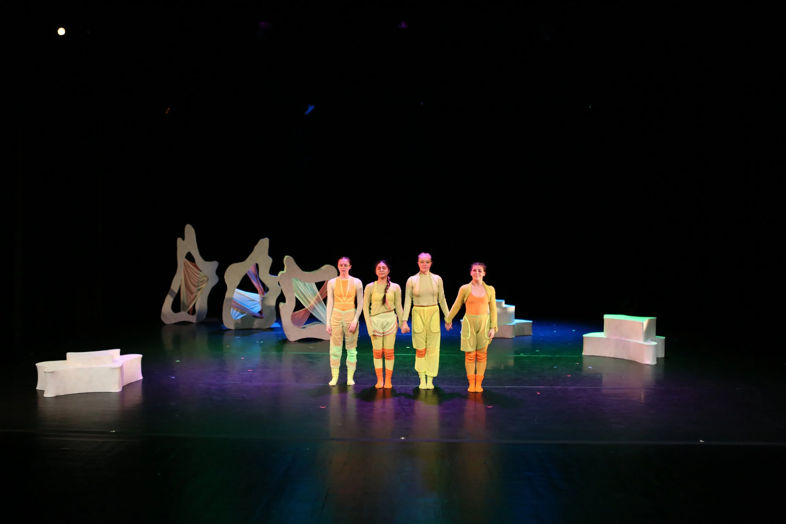 Four dancers holding hands in a line on stage with abstract set pieces in the background and minimal props on the stage.