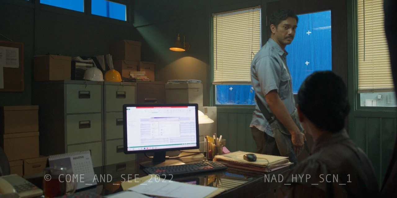 An office scene showing two men, one standing near two windows with closed blinds and the other seated at a desk. The desk has a computer monitor, files, and office supplies, with filing cabinets and boxes in the background.