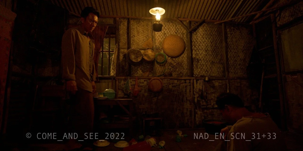 Two men in a dimly lit rustic room with woven walls, one standing and one sitting, surrounded by bowls and containers, with a single hanging light. 