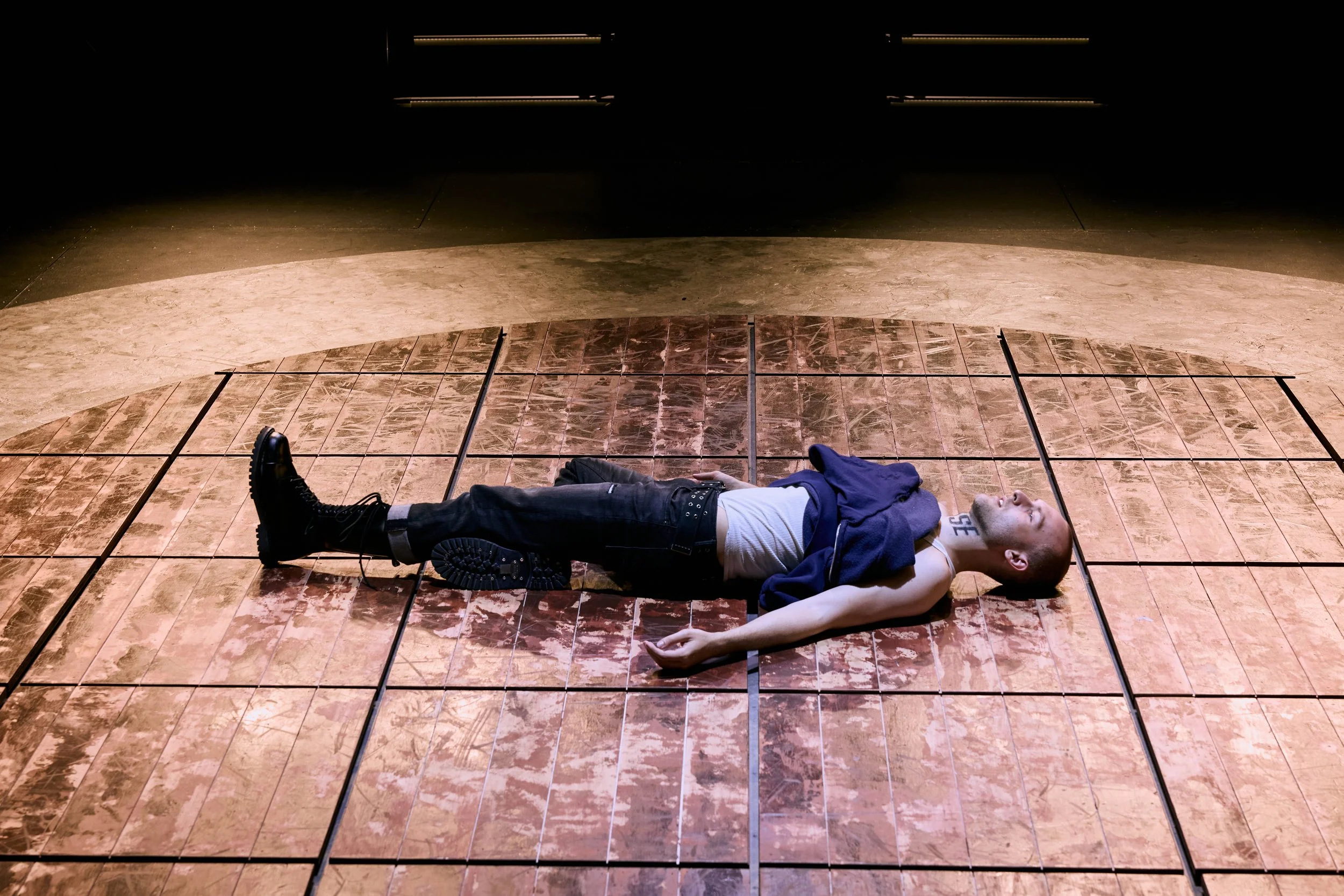 A person lying on a tiled floor in a room with black walls. They are wearing black boots, black pants, a white shirt, and a blue hoodie, with one arm extended on the floor. The room is dimly lit with lights on the ceiling.