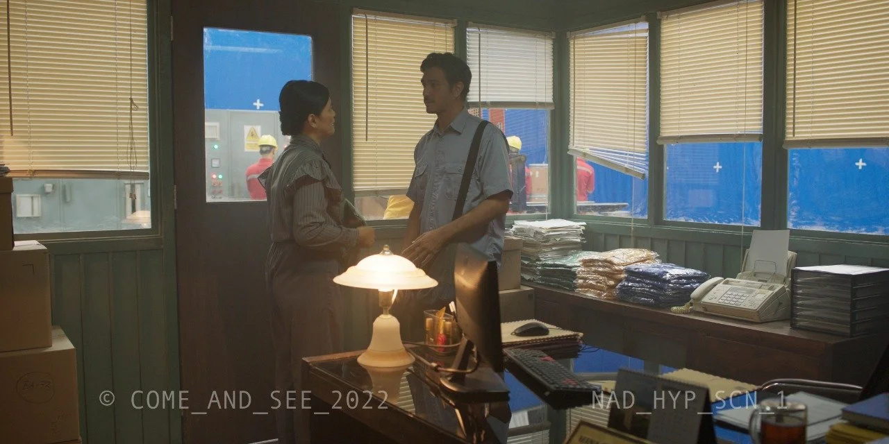 Two people talking in an office with a desk, computer, phone, and stacks of paper, while two workers in helmets work outside through the windows.