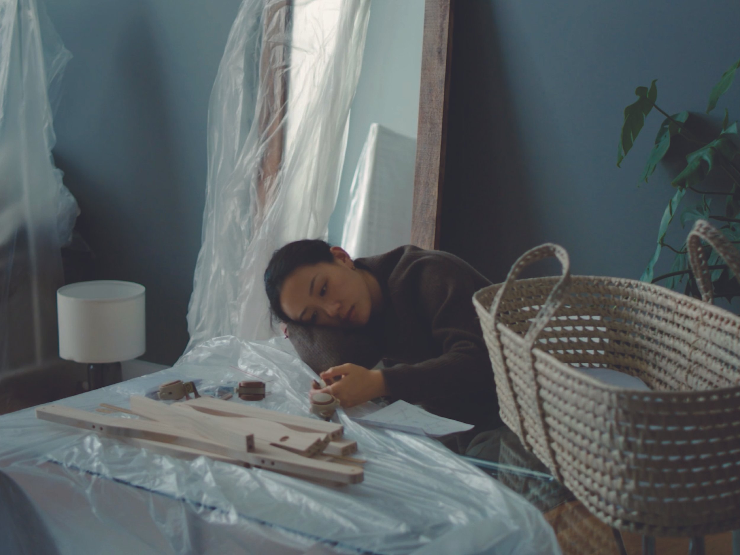 A person lying on a bed in a room with blue walls, next to a nightstand with a white lamp, surrounded by clear plastic sheets and some wooden objects, with a woven basket in the foreground and a potted plant in the background.