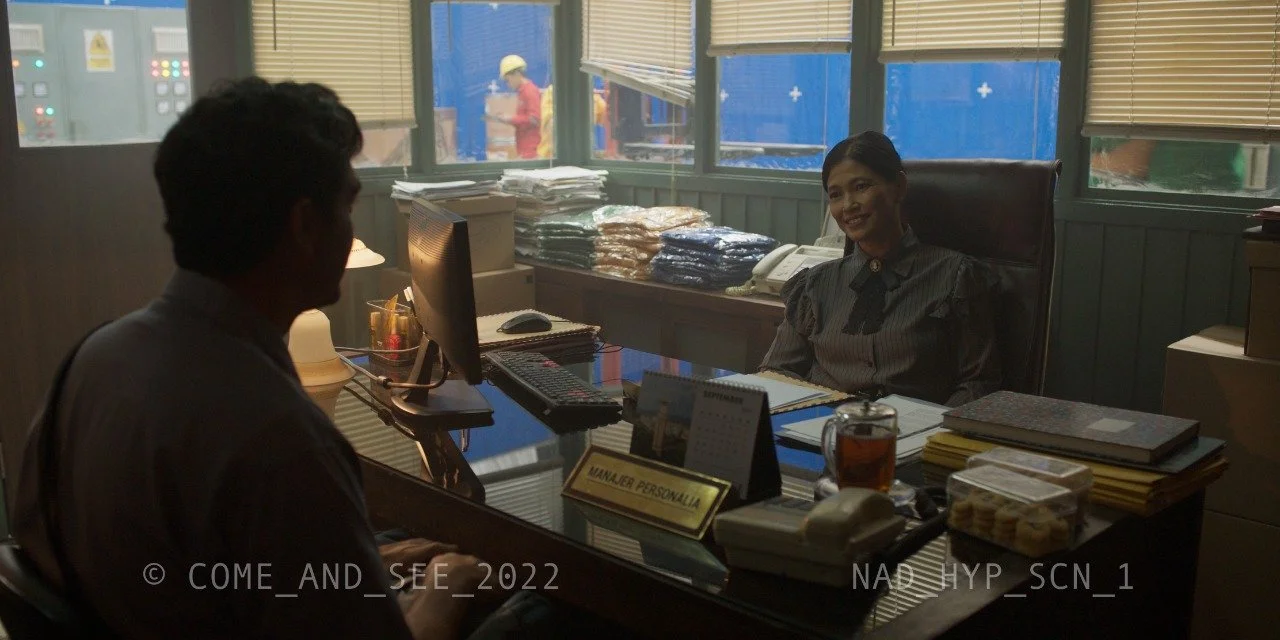 An office setting with two women. One woman with short hair is sitting at the desk, while the other woman, with longer hair, is sitting behind the desk. The desk has a computer, a nameplate, and various office supplies. Behind the woman sitting at th
