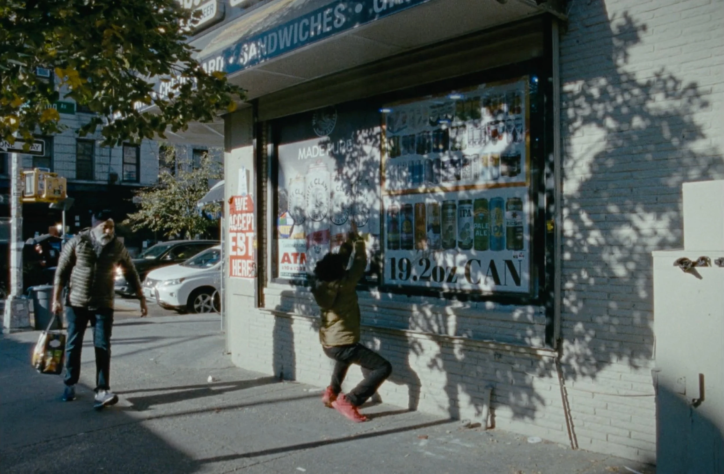 A sidewalk scene with two people, a woman and a child, walking past a closed store with a metal shutter. The store has a sign for beer and sandwiches, and inside the window are various posters and signs. Sunlight shadows cast leafy patterns on the bu