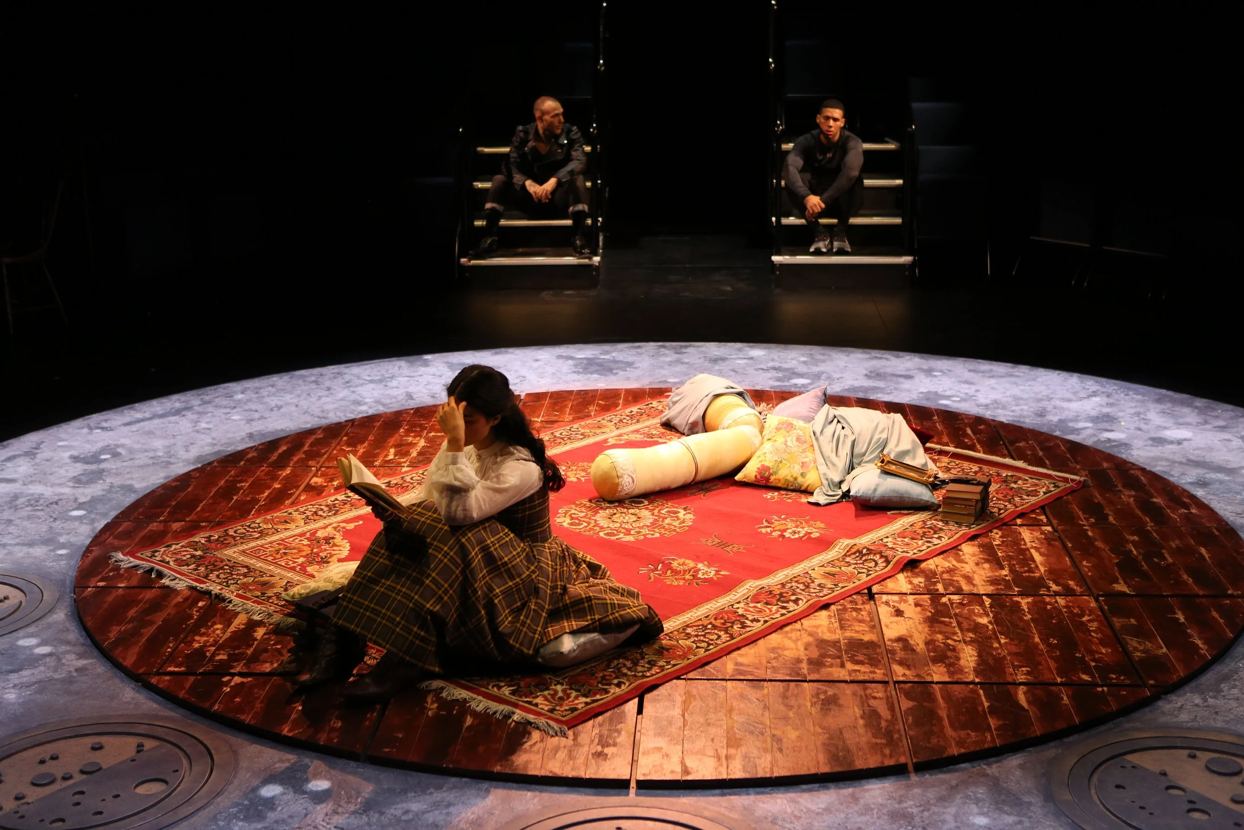 The image shows a theatrical stage with a circular rug, a woman sitting on the rug reading a book, and two men sitting on steps in the background. The stage is dimly lit.